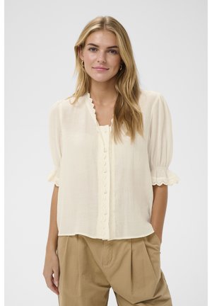 Giovane donna con lunghi capelli biondi che indossa una camicetta color crema con bordo smerlato e pantaloni beige a vita alta, in piedi davanti a uno sfondo semplice.