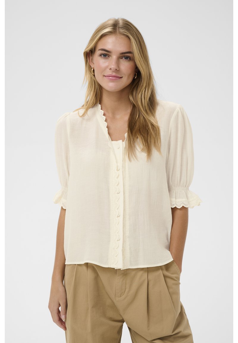 Noor naine pikkade blondide juustega kannab kreemikat pluusi lainelise äärega ja beeže kõrge vöökohaga pükse, seistes ühevärvilise tausta ees.