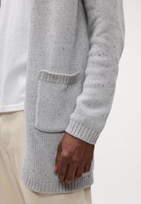 Gros plan d'une personne portant un cardigan tricoté gris clair avec une poche avant et des poignets côtelés, associé à un pantalon beige et une chemise blanche.