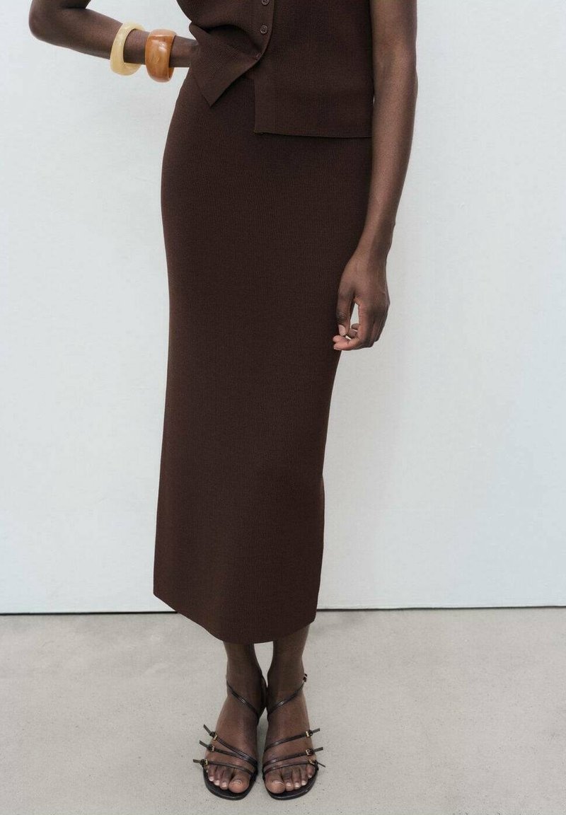 Femme portant une robe sans manches marron foncé avec des boutons, des bracelets en bois et des sandales noires à lanières, debout contre un mur uni.