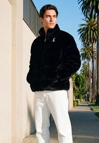 Muškarac u crnoj jakni od paperja i bijelim hlačama stoji na pločniku okruženom palmama u jarkom dnevnom svjetlu, ruke u džepovima jakne.