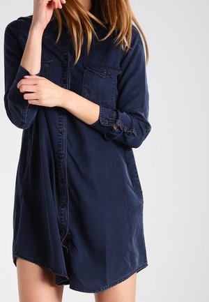 Femme portant une robe-chemise bleue foncée à manches longues avec des poches avant, ajustant sa manche d'une main.