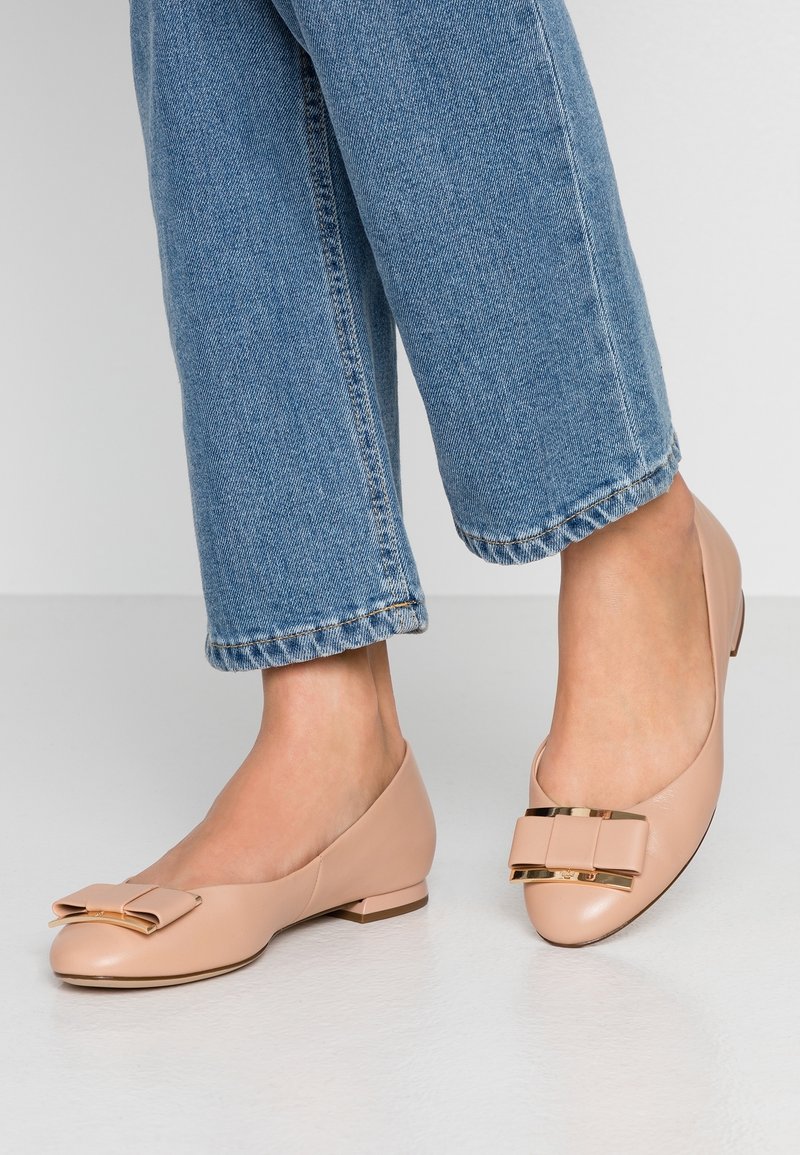 Pieds portant des ballerines rose clair avec un détail de noeud bordé d’or, associés à un jean bleu court sur un sol blanc.
