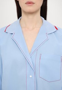 Femme portant une chemise bleu clair avec des coutures rouges et des accents rouges sur le col, montrant la partie supérieure de la poitrine et du cou.