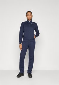 Femme debout avec une main dans la poche, portant une veste zippée Champion marine et un pantalon assorti, avec des chaussures de sport noires sur fond uni.