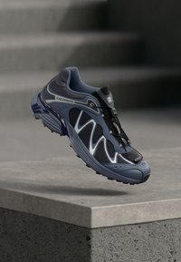 Αθλητικό παπούτσι Salomon σε σκούρο μπλε και γκρι, με επάνω μέρος από δίχτυ, μοναδικό κυματιστό σχέδιο και σύστημα κλειδώματος κορδονιών, διαθέτοντας υφή στη σόλα.