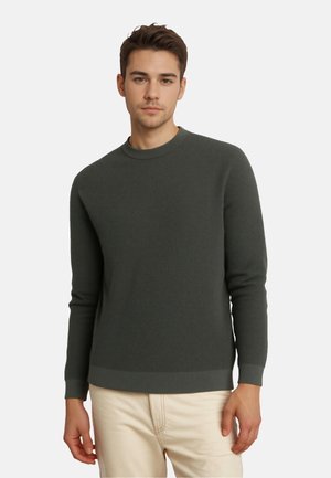 Pull côtelé vert foncé en tissu texturé, avec un col rond et des manches longues, associé à un pantalon de couleur claire.