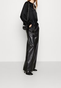 Donna che indossa una blusa di satin nera, pantaloni in pelle neri a gamba larga, stivali neri con tacco e una piccola borsa nera con tracolla a catena.