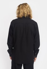 Camisa negra de manga larga con una tela texturizada, puños abotonados y dobladillo curvado. Detalle de yugo trasero visible.