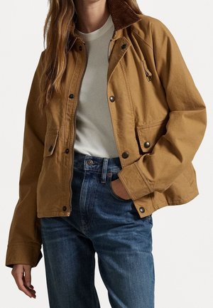 Persoon die een bruine toegeknoopte jas draagt over een wit overhemd en blauwe jeans, met één hand in de zak en de andere ontspannen langs het lichaam.