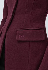 Close-up of a burgundy wool coat with a flap pocket featuring a stitched logo on the left side.