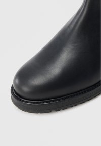 Černá kožená kotníková bota s hladkou texturou, kulatou špičkou a tlustou gumovou podrážkou s dezénem pro lepší přilnavost.