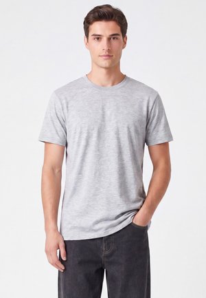 Giovane uomo con capelli corti castani che indossa una semplice t-shirt grigio chiaro e jeans scuri, in piedi con una mano in tasca.