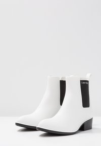 Botas blancas de tobillo hechas de material suave, con paneles elásticos laterales negros y un bajo tacón cuadrado. Diseño duradero y elegante con detalles de costura.