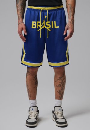 Osoba nosząca niebieskie spodenki do koszykówki z żółtym obszyciem i napisem „BRASIL”, w połączeniu z białymi i czarnymi sneakersami oraz białymi skarpetami.