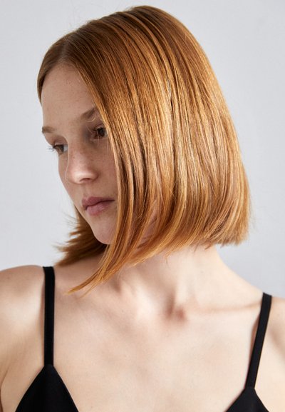 Short, straight copper hair; subtle texture. Black spaghetti-strap top. Neutral background; subject in profile looking downward.