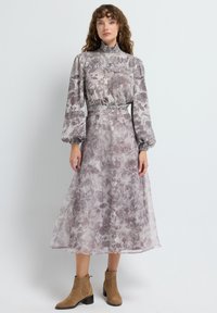 Mujer con un vestido midi floral de manga larga y cuello alto, con la cintura ceñida y botas tan hasta el tobillo, de pie frente a un fondo gris claro.