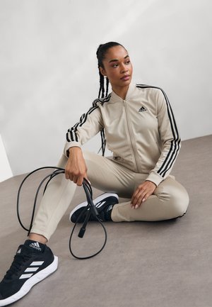 Tuta sportiva beige realizzata in tessuto liscio, con strisce nere. Scarpe da ginnastica nere con dettagli bianchi. Corda per saltare tenuta in mano.