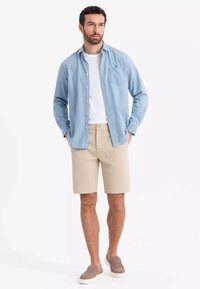Camicia button-up azzurro chiaro sopra una maglietta bianca, abbinata a pantaloni corti beige e scarpe slip-on marroni. Design semplice e vestibilità casual.