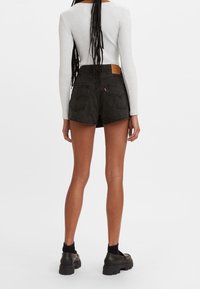 Kvinna med långa flätor, klädd i en vit ribbad långärmad topp, svarta högmidjade jeansshorts, svarta strumpor och kraftiga svarta skor, vänd bort.