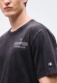 Νεαρός που φοράει ξεθωριασμένο μαύρο T-shirt Champion με το λογότυπο "Champion Training Club" στο στήθος και μικρό λογότυπο στο μανίκι.