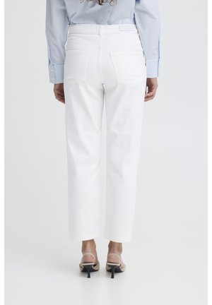 Žena stojaca chrbtom, oblečená v bielych nohaviciach s rovnými nohavicami, svetlomodrej košeli s dlhým rukávom a béžových sandáloch na podpätku, na bielom pozadí.