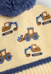 Gorro de punto en color crema con detalles en azul. Presenta tractores y excavadoras bordados en marrón, con un pompón azul esponjoso en la parte superior.