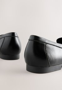 Paire de chaussures plates en cuir noir avec petits talons vues de dos sur un fond clair uni.