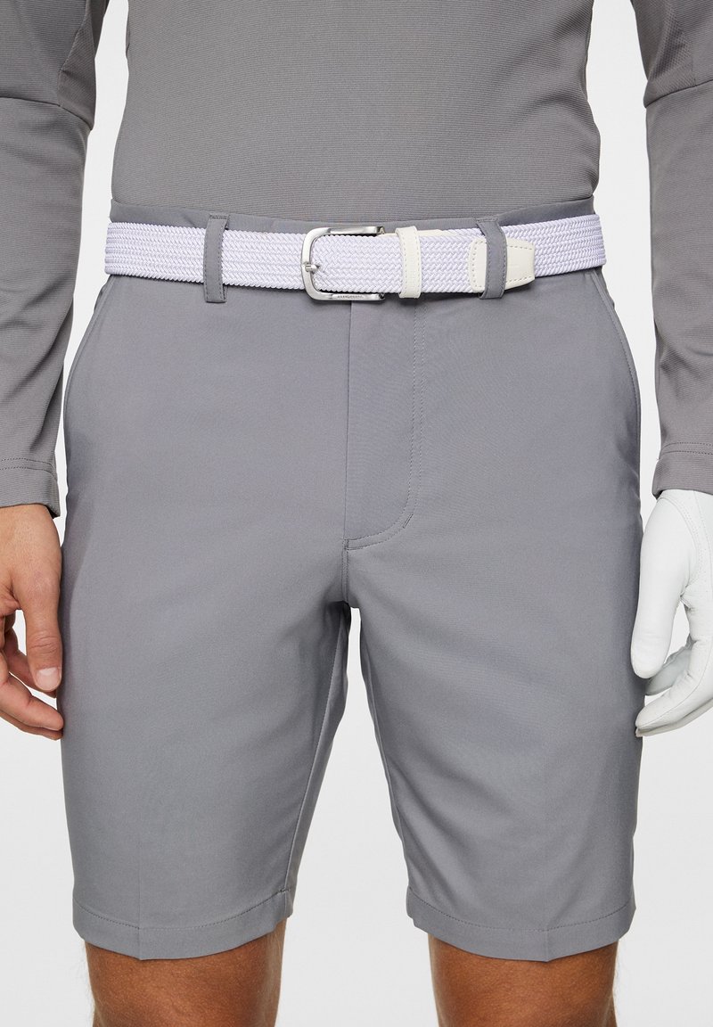 Harmaat golf-shortsit, joissa on hienovaraisia ommeltuja yksityiskohtia, yhdistettynä teksturoituun valkoiseen ja purppuraan punottuun vyöhön. Golf-käsine on vasemmassa kädessä.