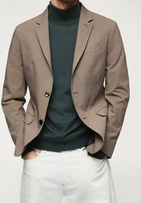 Ruskea räätälöity bleiseri, jossa on lovetut kaulukset, kaksi mustaa nappia ja rintatasku, käytetty tummanvihreän poolopaidan ja valkoisten housujen päällä.