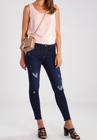 Top sin mangas rosa claro, jeans ajustados rasgados azul oscuro, sandalias negras de tacón con bordado floral. Atuendo casual con elementos de diseño moderno.