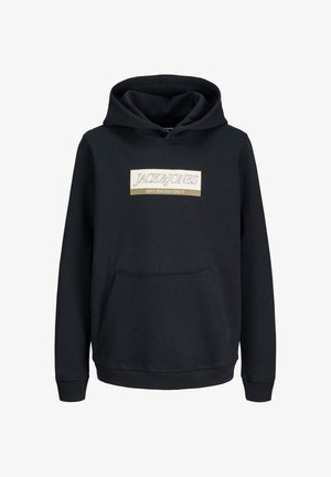Junge steht und lächelt, trägt einen schwarzen Hoodie mit Logo, dunkle Jeans und schwarze Sneakers vor einem schlichten hellen Hintergrund.