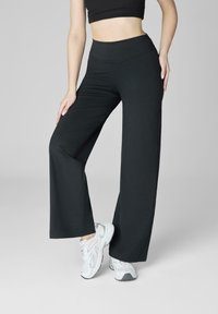 Kobieta ubrana w czarne, wysokie spodnie o szerokich nogawkach, czarny top crop i białe trampki, stojąca z jedną nogą lekko ugiętą.