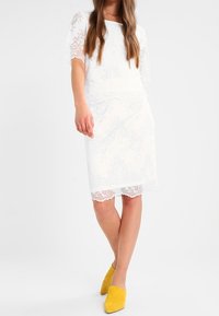 Weißes Spitzenkleid mit kurzen Ärmeln, verziert mit floralen Mustern und einem knielangen Saum. Kombiniert mit gelben High-Heels-Mules.