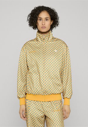 Person trägt eine gelbe und blaue Hahnentritt-RV-Jacke und passende Hose mit Logos auf der Brust und dem Oberschenkel, steht aufrecht.