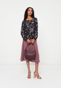 Langärmlige schwarze Bluse mit lila und grauem abstraktem Muster, kombiniert mit einem fließenden mauvefarbenen Rock und einer burgunderfarbenen Umhängetasche. Beige Blockabsatzsandalen.