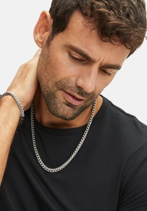 Silver metal chain necklace with interlocking links, worn over a black t-shirt. Close-up of textured design and smooth finish.