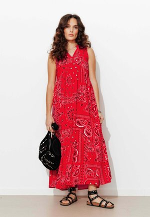 Femme aux longs cheveux bouclés portant une robe longue sans manches rouge avec un imprimé paisley, tenant un sac noir clouté et portant des sandales noires cloutées.