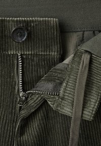 Pantalones de pana verde oliva con un botón negro, cremallera de metal y ribete de tela. La superficie tiene una textura acanalada y acentos suaves.