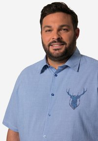Camisa de mangas cortas a cuadros azul y blanca con collar extendido, que presenta un diseño de ciervo azul en el lado izquierdo del pecho y botones azules.