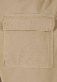 Poche en tissu beige avec rabat cousue sur un tissu beige similaire, détaillée avec des coutures doubles le long des bords.