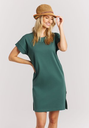 Vrouw met een groene jurk met korte mouwen en een geweven strohoed, glimlachend met gesloten ogen, de ene hand op haar heup en de andere om de hoed recht te zetten.