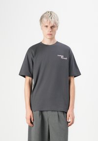 Junge Person mit kurzen hellen Haaren, die ein dunkelgraues T-Shirt mit dem Text "Trattoria Remo's" und lockeren grauen Hosen vor einem einfarbigen Hintergrund trägt.