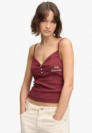 Jeune femme aux cheveux bruns courts portant un haut sans manches bordeaux avec des boutons et un pantalon blanc, debout les mains dans les poches.