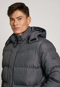 Siva puffer jakna s kapuljačom, izrađena od teksturiranog materijala, s prednjim zatvaračem i čičak trakama na ovratniku i ramenu.