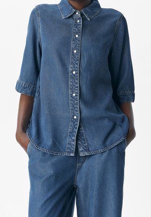 Femme portant une chemise en jean bleue à boutons avec manches retroussées et un pantalon ample en jean assorti, mains dans les poches.
