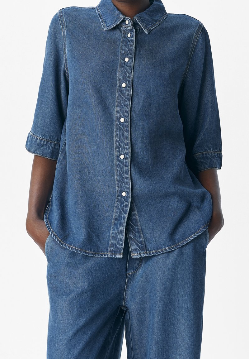 Femme portant une chemise en jean bleue à boutons avec manches retroussées et un pantalon ample en jean assorti, mains dans les poches.