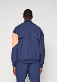 Marineblaue Sportjacke mit einem orangefarbenen Akzent, ausgestattet mit hohem Kragen, elastischen Bündchen und Details am Rückenschlitz. Glattes, leichtes Material.