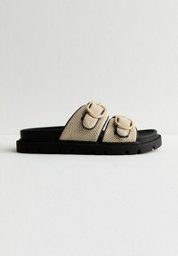 Beige vävd halksandaler med två gyllene spännen, svart flexibel sula och texturerad ovansida. Storlek och varumärke syns inte.
