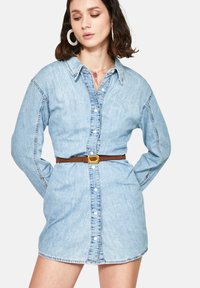 Vestido camisero de denim en azul claro, con botones delanteros, mangas largas y un cinturón marrón con hebilla cuadrada dorada en la cintura.